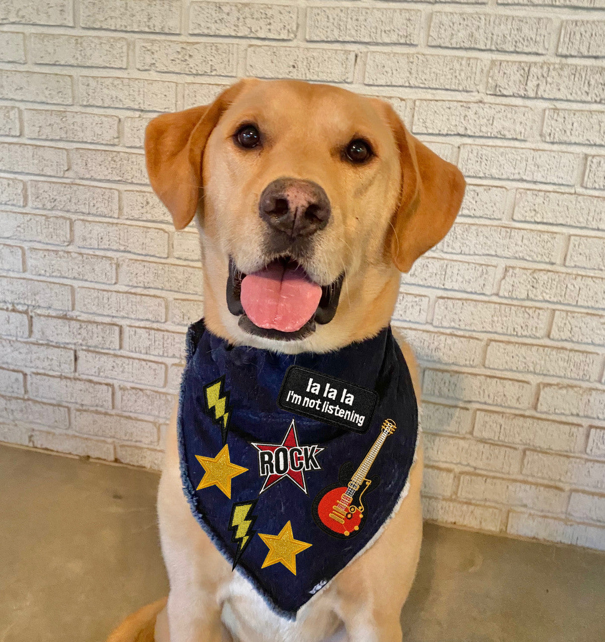 Happy Labrador retriever wearing a custom denim bandana with rock and roll patches, sitting in front of a brick wall